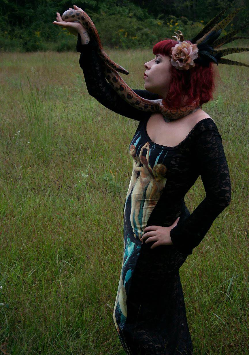 A slender redheaded model wearing a Three Graces print dress holds a Brazilian Rainbow Boa named Mercury.